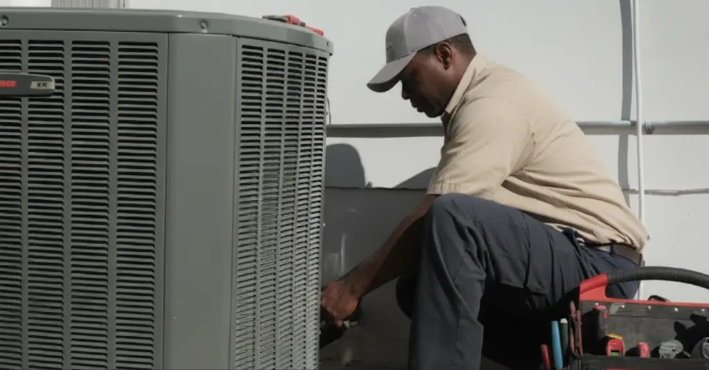 An HVAC service contractor kneels on the ground next to the outdoor air conditioning unit he is servicing.