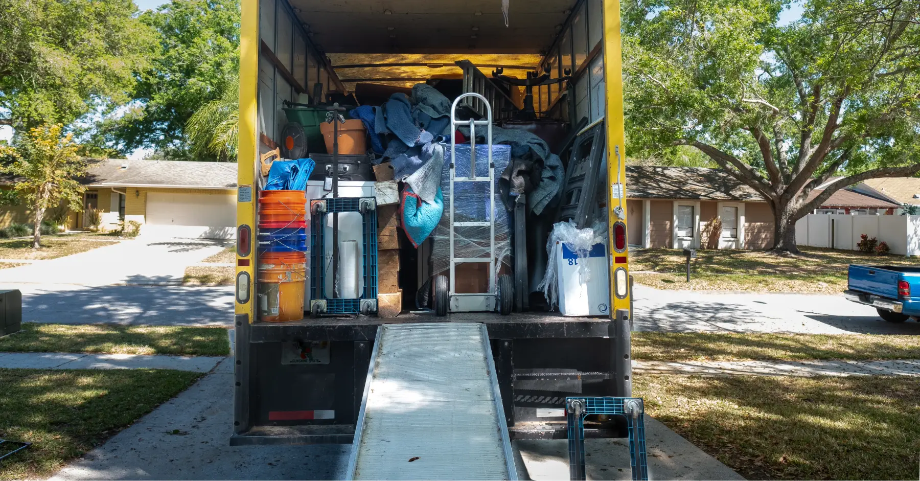 A moving truck is parked in the driveway of a home with the ramp down. The back door is open and the items in the truck are stacked inside, ready to be moved.